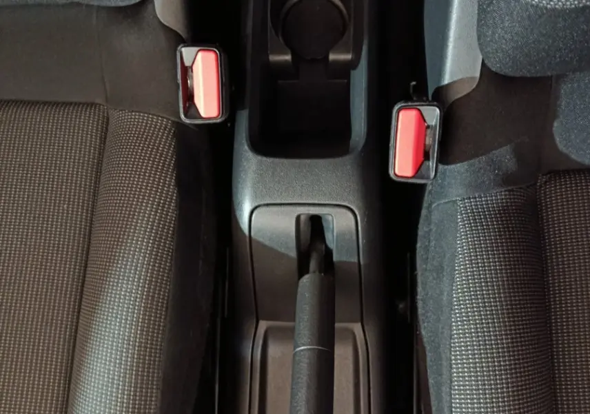 Vue plongeante sur la console centrale noire entre les sièges avant gris à motifs, avec frein à main et boucles de ceinture rouges visibles.