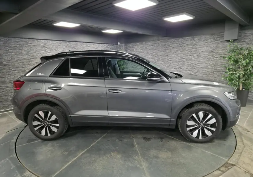 Vue latérale droite d'un Volkswagen T-Roc gris métallisé édition GOAL avec toit et montants noirs en intérieur showroom.