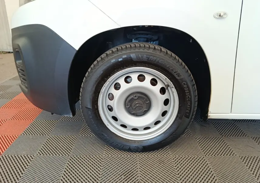 Gros plan sur la roue avant gauche en jante acier du Citroën Berlingo blanc, avec partie basse du pare-chocs noir visible.