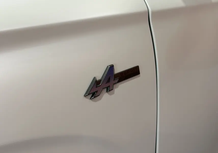 Gros plan sur l'écusson Esprit Alpine violet sur la porte blanche nacrée satinée d'un Renault Austral.