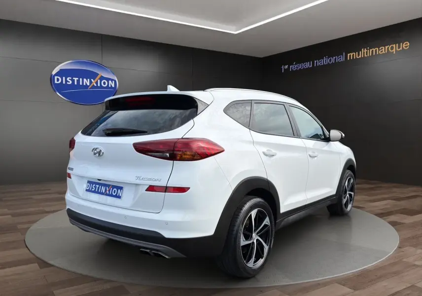 Vue 3/4 arrière droite d'un Hyundai Tucson blanc polar avec jantes 18 pouces bicolores, sur plateforme en showroom.