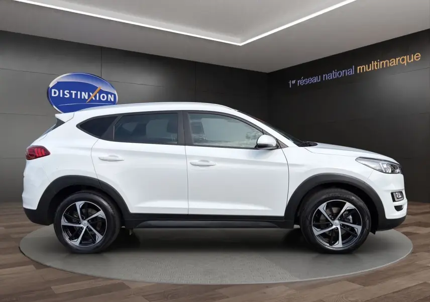 Vue de profil côté gauche d'un Hyundai Tucson blanc polar avec jantes 18 pouces bicolores dans un showroom.