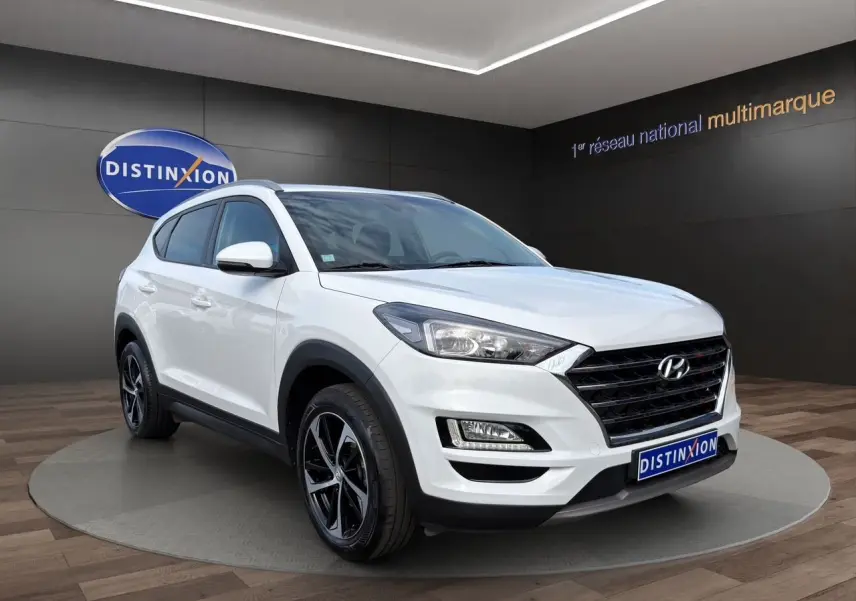 Hyundai Tucson blanc polar en 3/4 avant droit, avec jantes 18 pouces bi-ton et calandre noire distinctive.