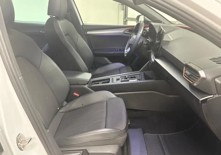 Vue intérieure côté conducteur de la CUPRA Leon 2024 blanche Nevada, avec sièges noirs et tableau de bord moderne.