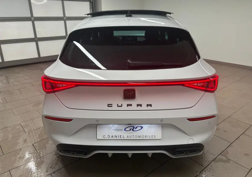 Vue arrière d'une CUPRA Leon blanche Nevada avec feux arrière allumés et toit ouvrant panoramique visible.