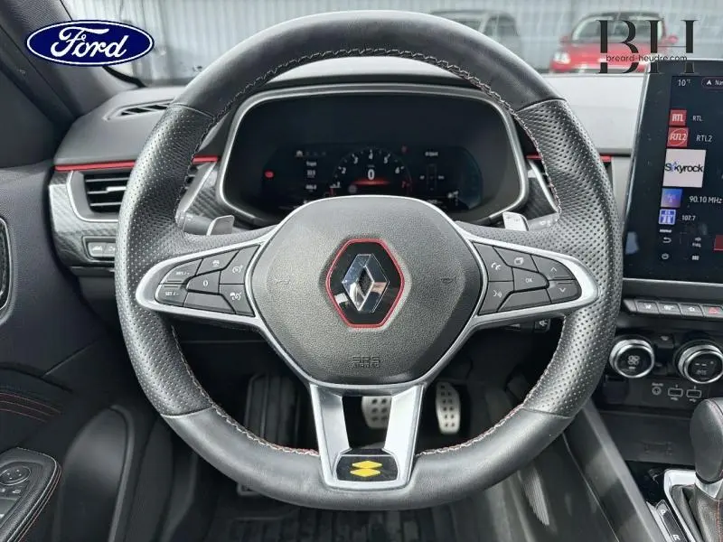 Vue rapprochée du volant cuir noir avec surpiqûres rouges et logo Renault au centre d’un Renault Arkana gris métallisé.