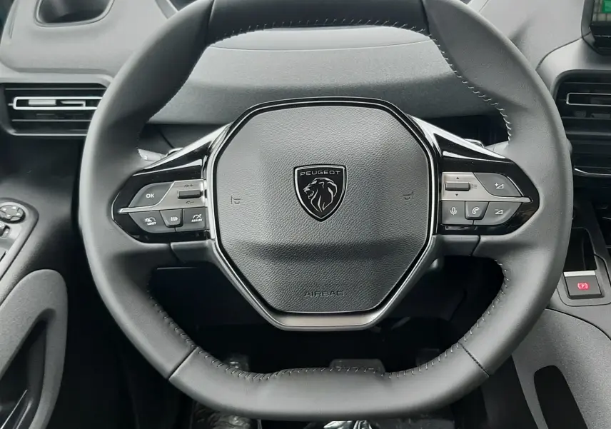 Vue rapprochée du volant noir du Peugeot Rifter 2026 avec commandes intégrées et tableau de bord numérique en arrière-plan.