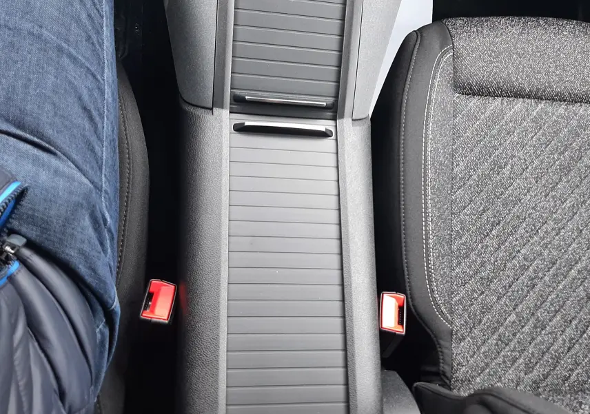 Vue plongeante sur la console centrale noire et les sièges tissu gris du Peugeot Rifter, avec ceinture rouge visible.