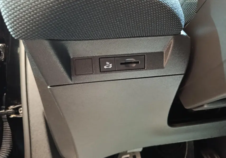 Gros plan sur le bouton d'ouverture du coffre noir situé sous le siège avant du Citroën C5 Hybride 2026.