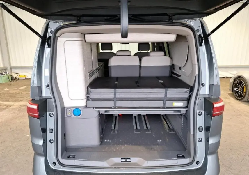 Vue arrière du Volkswagen California T7 gris Indium avec hayon ouvert montrant l'intérieur modulable et la couchette repliée.