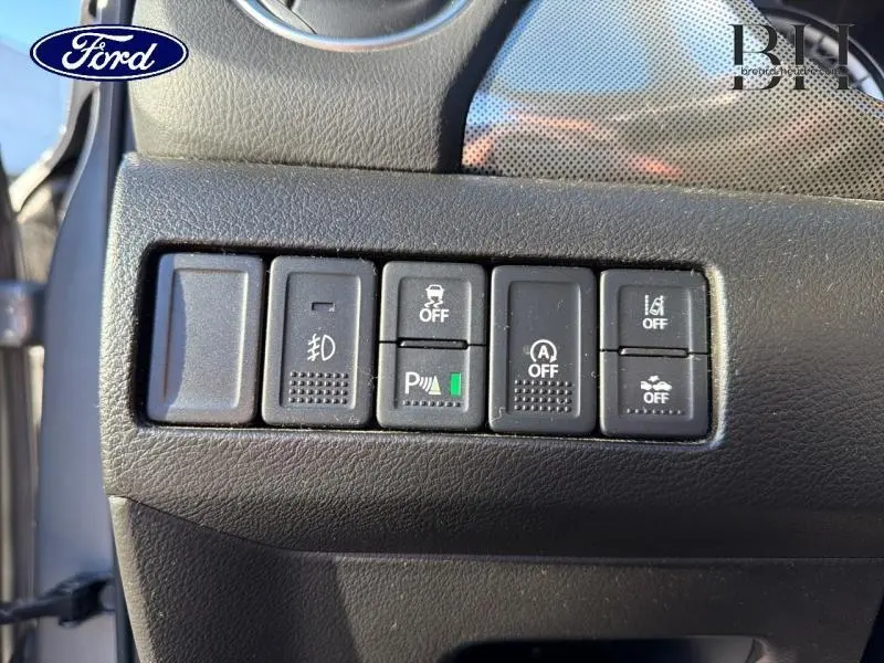 Gros plan sur les boutons de commande noirs du tableau de bord côté gauche, avec icônes de sécurité et assistance, intérieur Ford.
