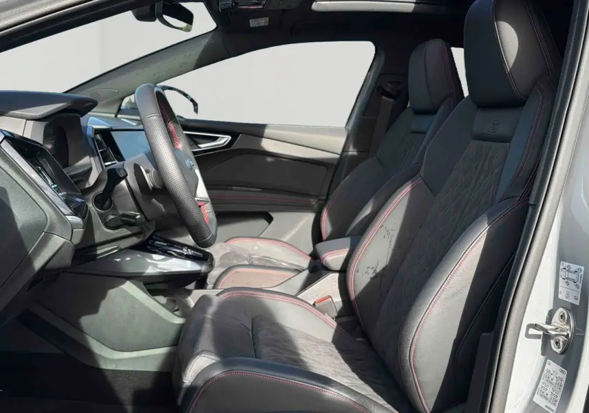 Vue intérieure côté gauche de l'Audi Q4 e-tron 2025, sièges en cuir noir avec surpiqûres rouges et volant multifonction.