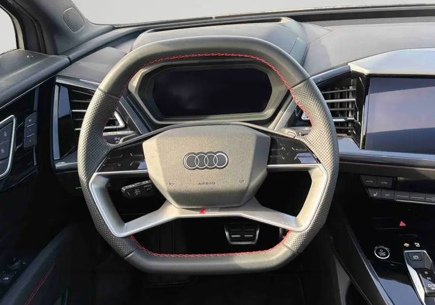 Volant multifonction noir avec surpiqûres rouges dans l'habitacle d'un Audi Q4 e-tron 2025, tableau de bord et écran tactile visibles.