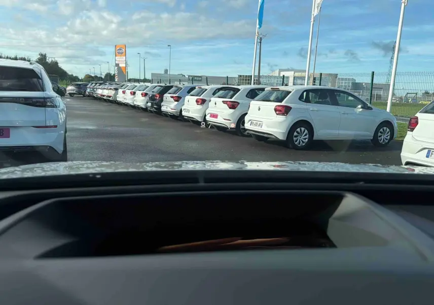 Vue depuis l'intérieur montrant le tableau de bord et plusieurs Volkswagen blanches garées à l'extérieur.