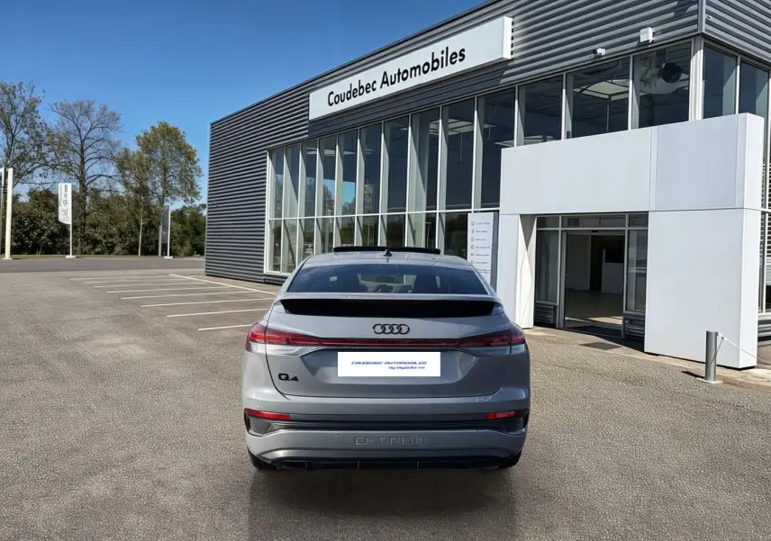 Vue arrière d'un Audi Q4 e-tron gris galet stationné devant un bâtiment moderne de concession automobile.