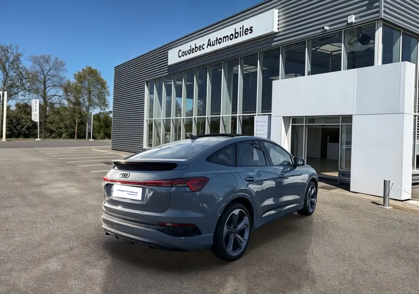 Audi Q4 e-tron gris galet en 3/4 arrière droit, avec jantes 19 pouces, stationné devant un bâtiment moderne.