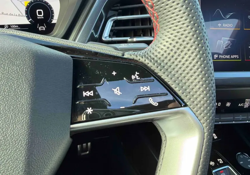 Gros plan sur le volant en cuir perforé avec commandes tactiles à droite, tableau de bord numérique et écran central visibles.