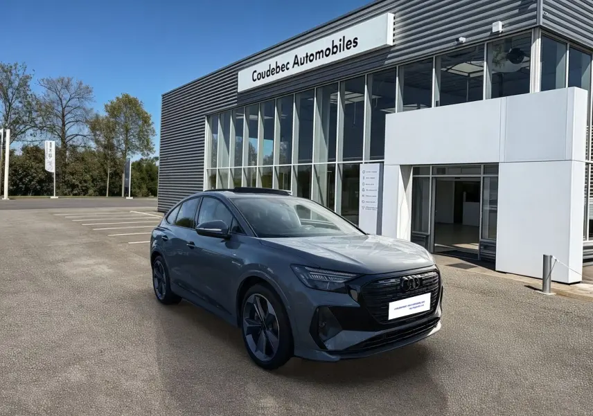 Audi Q4 e-tron gris galet en 3/4 avant droit devant un bâtiment de concession Coudebec Automobiles.