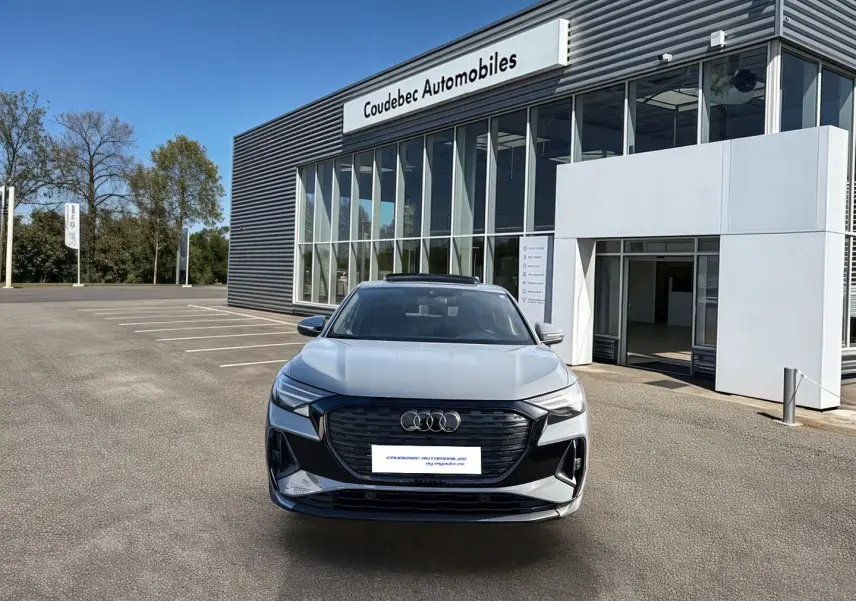 Audi Q4 e-tron gris galet vu de face, stationné devant un bâtiment moderne avec large baie vitrée.