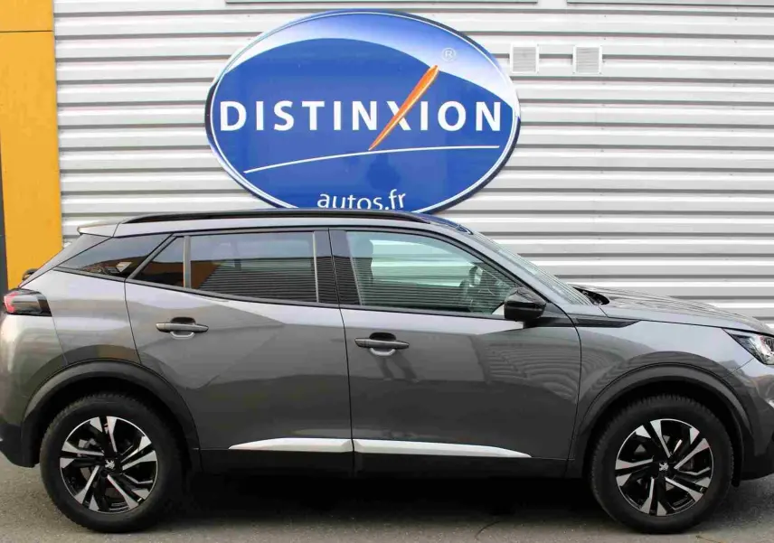 Profil droit d'un Peugeot 2008 gris platinium avec jantes noires et toit panoramique visible, devant un mur métallique.