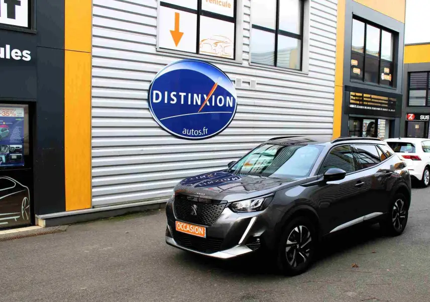 Peugeot 2008 gris platinium en 3/4 avant droit, avec calandre noire et toit panoramique visible devant un bâtiment commercial.
