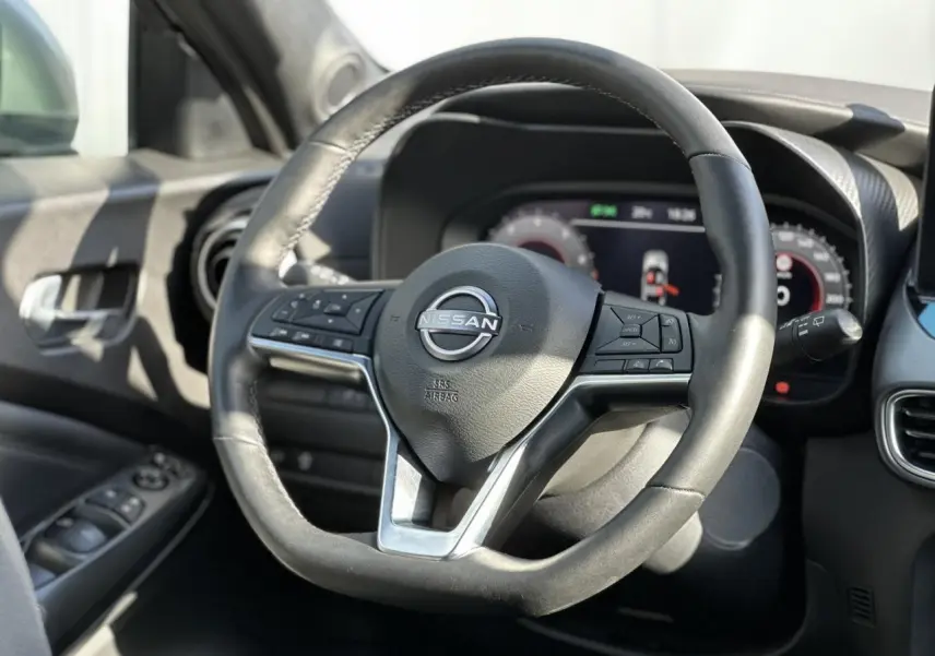 Vue rapprochée du volant en cuir noir du Nissan Juke 2025 avec commandes intégrées et tableau de bord digital en arrière-plan.