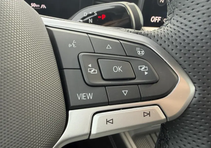 Gros plan sur les commandes noires et argent du volant perforé du Volkswagen T-Roc 2026, avec affichage numérique en arrière-plan.