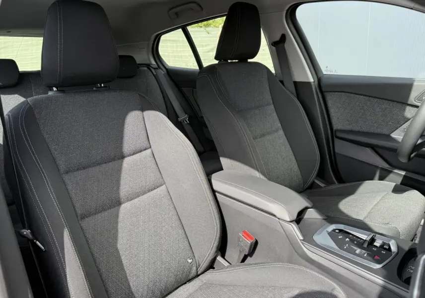 Vue intérieure côté avant droit de la BMW Série 1 118d M Sport 2025, sièges tissu anthracite avec accoudoir central.