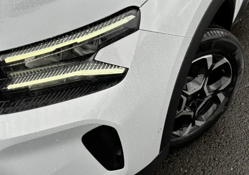 Gros plan sur le phare avant gauche et la roue alliage bi-ton noir du Citroën C5 Aircross blanc Okénite sous la pluie.