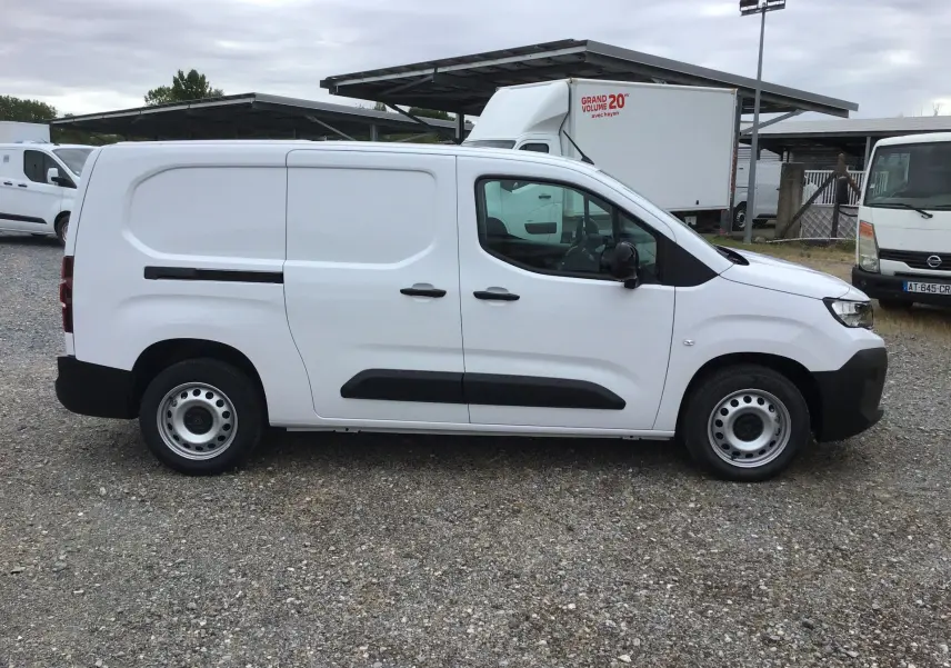Vue de profil côté gauche d’un Peugeot Partner blanc utilitaire avec protections noires et jantes acier, stationné sur sol gravillonné.