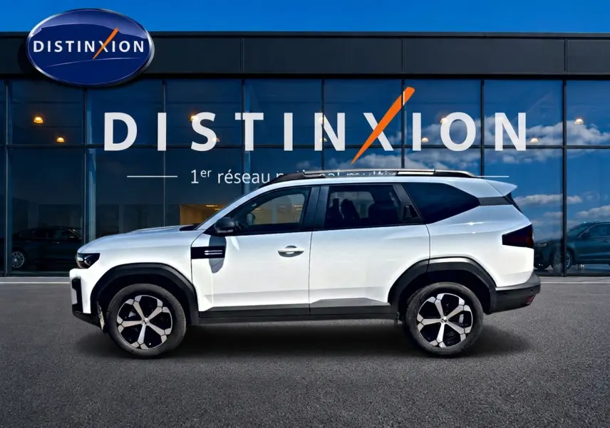 Vue de profil côté gauche d'un Dacia Bigster blanc avec jantes alu et barres de toit devant une vitrine Distinxion.
