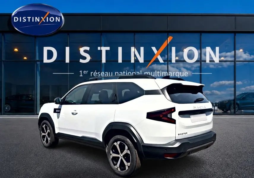 Vue 3/4 arrière droite du Dacia Bigster blanc avec jantes alu et feux arrière en forme de Z devant une vitrine Distinxion.