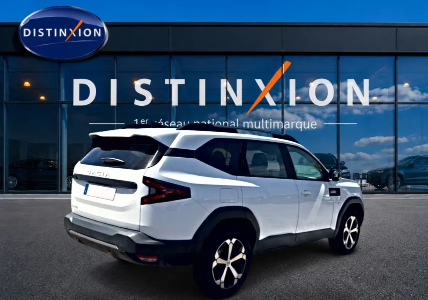 Vue 3/4 arrière droite du SUV blanc Dacia Bigster 2026 avec jantes alu et barres de toit noires devant un bâtiment Distinxion.