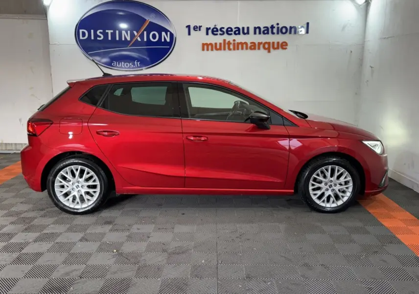 Profil latéral droit d'une SEAT Ibiza 1.0 TSI 110 FR rouge 2024, stationnée en intérieur sur sol damier noir et orange.