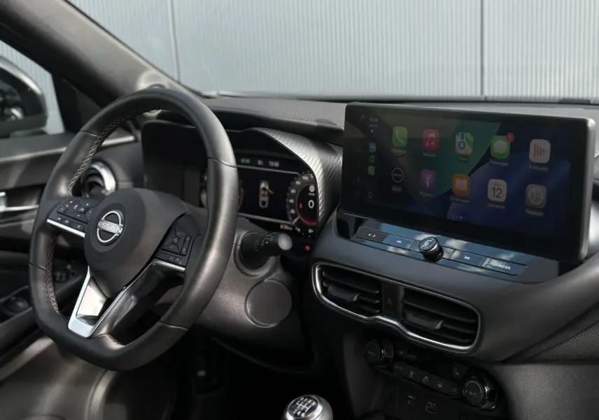 Vue intérieure côté conducteur du Nissan Juke 2025 gris foncé, volant cuir, écran tactile et levier de vitesse manuel.