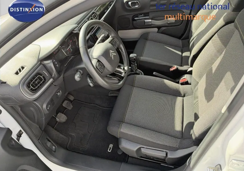 Vue intérieure côté conducteur de la Citroën C3 blanche 2021, montrant le volant, les sièges tissu gris et la boîte manuelle.
