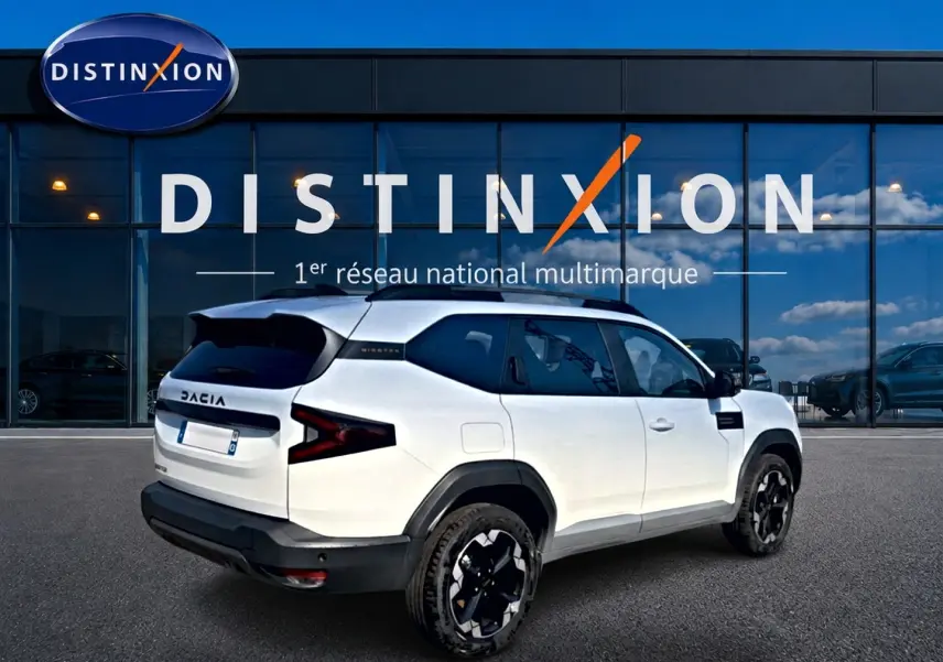 Vue 3/4 arrière droit du Dacia Bigster blanc avec jantes noires et toit noir devant une vitrine Distinxion.