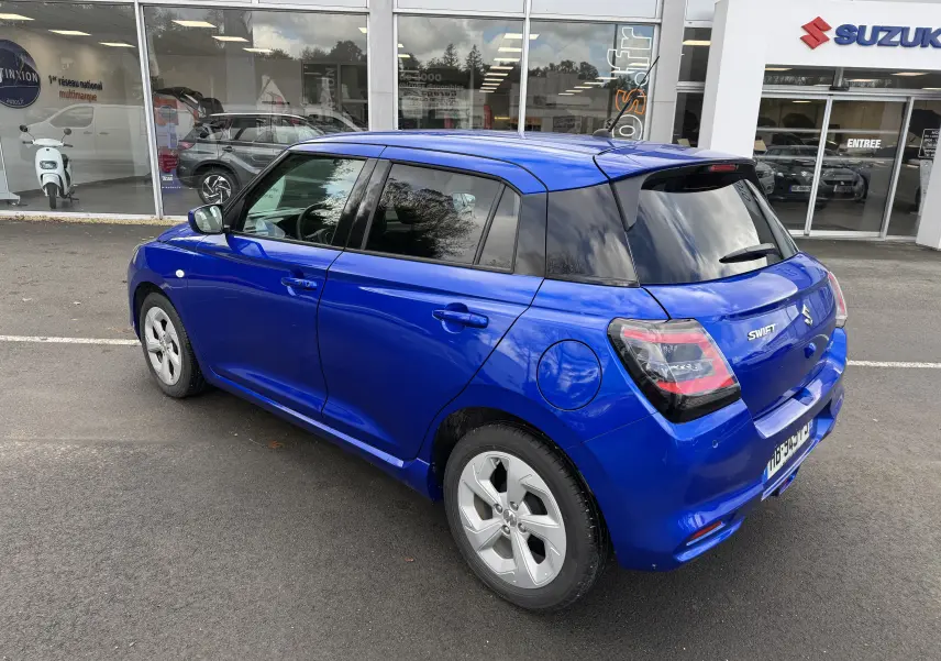 Vue 3/4 arrière droite d'une Suzuki Swift 1.2 Hybrid Privilège bleu, avec vitres teintées et jantes spécifiques, devant un concessionnaire.