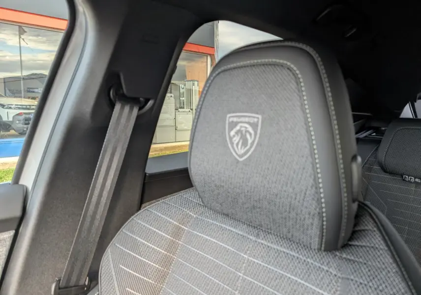 Gros plan sur l'appui-tête avant gauche gris avec logo Peugeot, intérieur tissu et ceinture de sécurité visible.