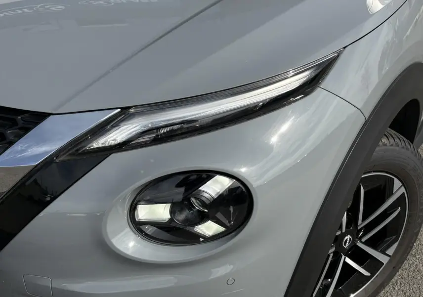 Gros plan sur le phare avant droit et la roue d’une Nissan Juke gris Ceramic, mettant en valeur les détails lumineux et la jante noire.