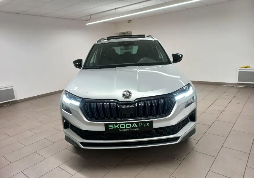 Vue avant d'un Skoda Karoq gris argent 2025 avec phares allumés et toit panoramique ouvrant visible en intérieur.