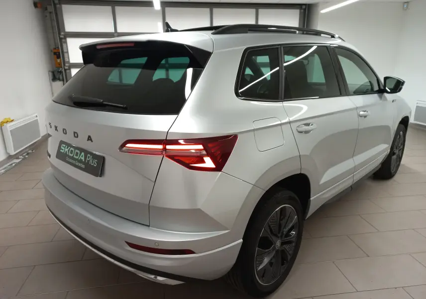 Vue 3/4 arrière droite du Skoda Karoq gris argent 2025 avec toit panoramique et feux arrière LED allumés.