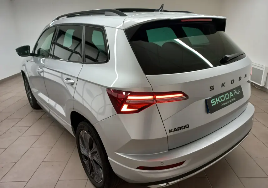 Vue 3/4 arrière droite du Skoda Karoq gris argent 2025 avec feux allumés et toit panoramique ouvrant.