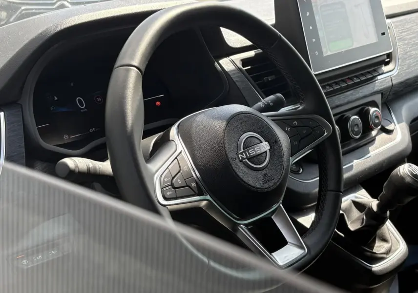 Vue rapprochée du volant et tableau de bord noir du Nissan Primastar Combi avec levier de vitesses manuel et écran tactile.