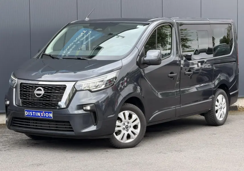 Nissan Primastar gris comète en vue 3/4 avant droit, avec hayon vitré et vitres latérales teintées.