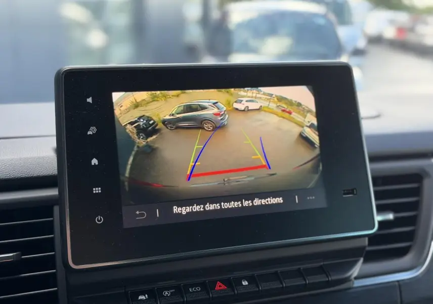 Écran de caméra de recul affichant un parking avec plusieurs véhicules, vu depuis l’habitacle du Nissan Primastar gris.