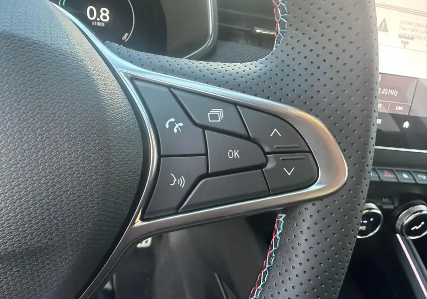 Gros plan sur les commandes multifonctions à gauche du volant perforé noir avec surpiqûres tricolores d’une Renault Clio V rouge.