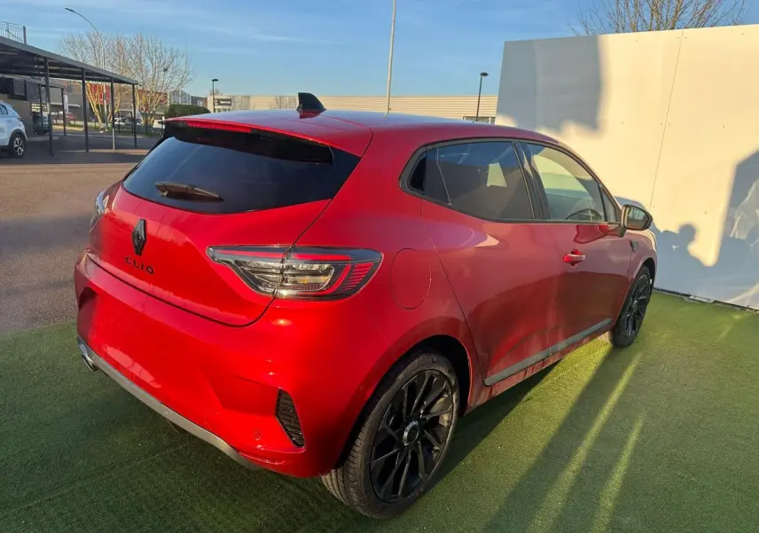 Vue 3/4 arrière droite d'une Renault Clio rouge foncé avec jantes noires et vitres teintées sur parking extérieur.