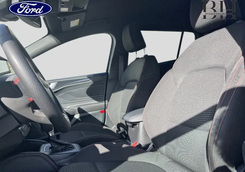 Intérieur avant de la Ford Focus SW 2024, sièges tissu noir avec surpiqûres rouges et volant cuir perforé.