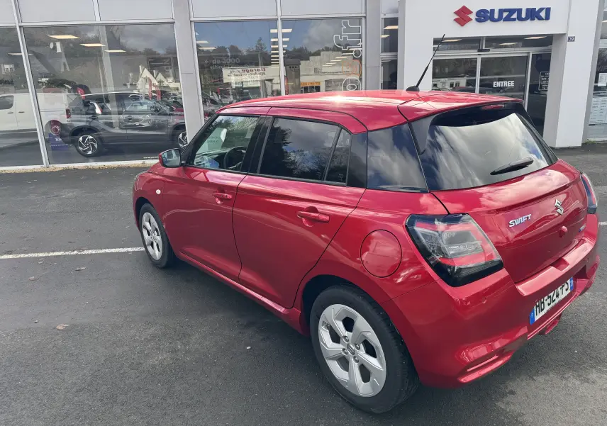 Vue 3/4 arrière droite d'une Suzuki Swift 1.2 Hybrid Privilege 2025 en rouge Burning Pearl, stationnée devant un concessionnaire.